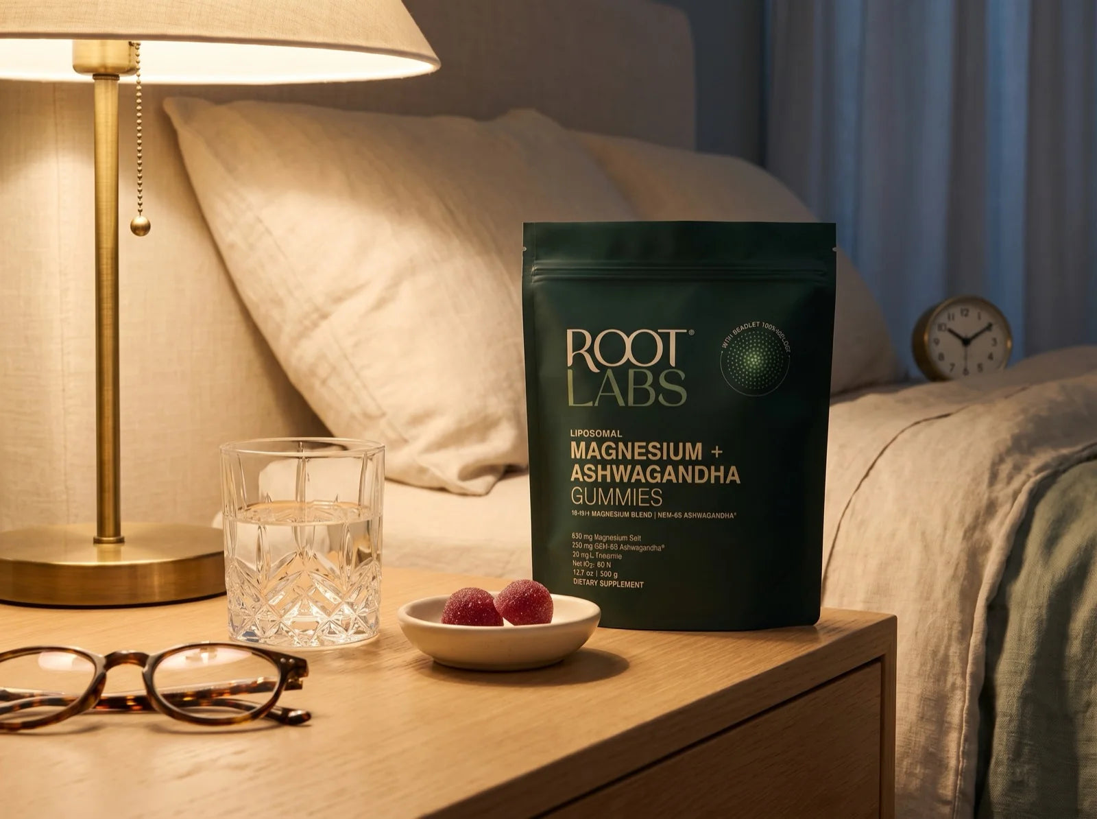 Root Labs AshwaMag pouch on a white oak nightstand at night: two berry gummies in a small ceramic dish, a cut-glass water tumbler, folded reading glasses, and a warm brass lamp